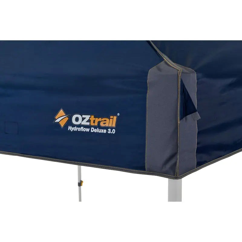 Close-up of the navy blue gazebo canopy corner featuring the white and orange OZtrail Hydroflow Deluxe 3.0 branding.
