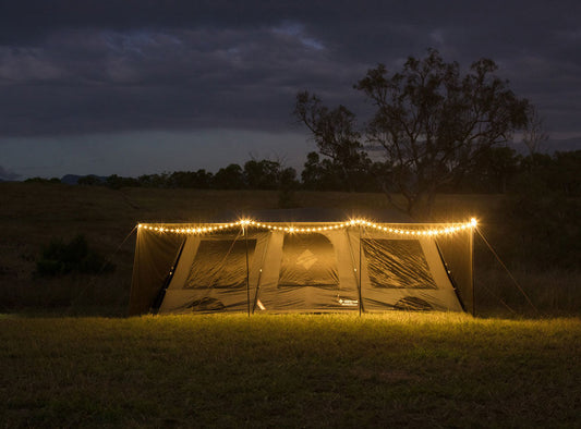 A string of glowing OZtrail Lumos fairy lights used to decorate the front awning of a large family tent at night in a grassy field.
